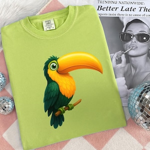 May include: A light green t-shirt featuring a colorful toucan illustration. The toucan has a large yellow beak, orange chest, and green and blue feathers. The shirt has a crew neck and a small tag on the collar.
