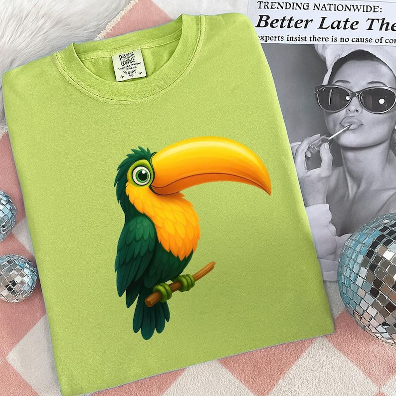May include: A light green t-shirt featuring a colorful toucan illustration. The toucan has a large yellow beak, orange chest, and green and blue feathers. The shirt has a crew neck and a small tag on the collar.