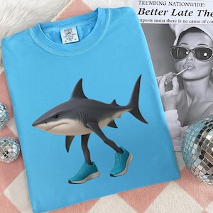 May include: A light blue t-shirt featuring a shark with legs and blue sneakers. The shark is gray and white. The shirt has a tag on the upper left side. The shirt is on a pink and white patterned surface.