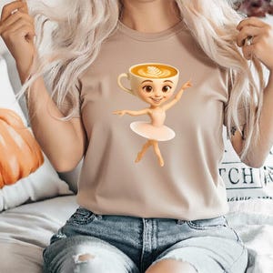 May include: A tan t-shirt featuring a cartoon ballerina with a coffee cup for a head. The ballerina wears a pink tutu and ballet shoes. The coffee cup has a latte art design.