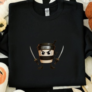 May include: Black sweatshirt featuring a cartoon coffee cup ninja character with a black headband, holding two silver katanas. The coffee cup has large eyes and is wearing a black ninja outfit. The design is centered on the front of the sweatshirt.