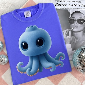May include: A blue t-shirt featuring a cartoon octopus with large eyes and tentacles. The octopus is light blue with darker blue accents and orange details. The shirt has a crew neck and a small tag on the upper left.