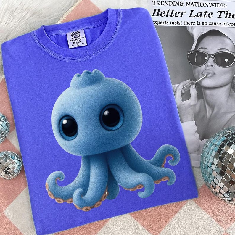 May include: A blue t-shirt featuring a cartoon octopus with large eyes and tentacles. The octopus is light blue with darker blue accents and orange details. The shirt has a crew neck and a small tag on the upper left.