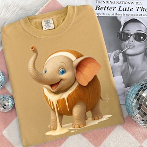 May include: Mustard-colored t-shirt featuring a cartoon elephant with a cream-colored design. The elephant has blue eyes and orange ears. The shirt has a crew neck and short sleeves. The tag reads "The Inspire Company".