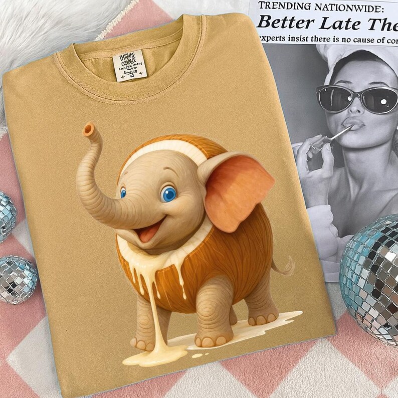 May include: Mustard-colored t-shirt featuring a cartoon elephant with a cream-colored design. The elephant has blue eyes and orange ears. The shirt has a crew neck and short sleeves. The tag reads "The Inspire Company".