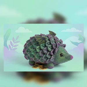 Op de afbeelding: Een handgemaakte gehaakte draak-egel hybride in groen en paars, met kleine vleugels en een roze neus. Het wezen heeft een getextureerd lichaam en wordt vergezeld door gouden munten, wat een fantasiethema suggereert.