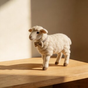 Op de afbeelding: Handgemaakte, off-white vilten schapenfiguur met een kleine strik en kraag. Het schaap heeft bruine hoeven en staat op een houten oppervlak. Een charmant decoratief item.