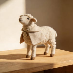 May include: A handcrafted, off-white sheep figurine with a cream-colored cape and a small, round pendant. The sheep has a fluffy, textured body, brown hooves, and is posed looking upwards. The figurine is displayed on a wooden surface.