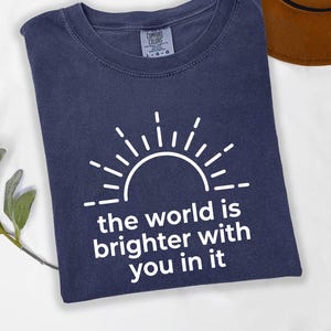 May include: A navy blue short-sleeved t-shirt featuring a white graphic of a sun rising above the text "the world is brighter with you in it". The shirt has a crew neckline and a visible brand label on the inside collar area.
