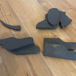 May include: Various grey wooden dog-shaped cutouts are displayed on a wooden surface. The set includes a head, ears, body, and tail, all separate. These appear to be components for a craft or decorative item. The wood shows a natural grain.