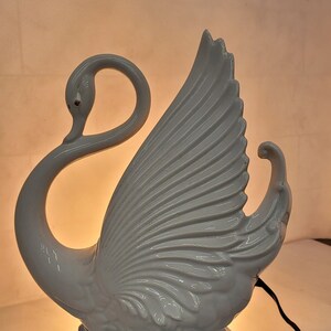 May include: A white ceramic swan lamp with a red eye detail. The swan has a curved neck and outstretched wing, with a light blue base. The lamp is lit from within, casting a warm glow.