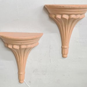 May include: A pair of peach-colored wall-mounted shelves featuring a semi-circular top surface and a decorative, fluted support bracket that tapers downward to a rounded base. The shelves have a smooth finish and a classic architectural design.