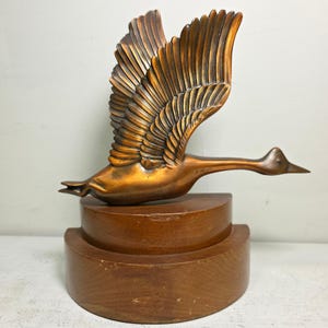 May include: A bronze-colored sculpture of a goose in flight, wings fully extended. The bird is mounted on a two-tiered, semi-circular wooden base. The sculpture has a detailed feather design.