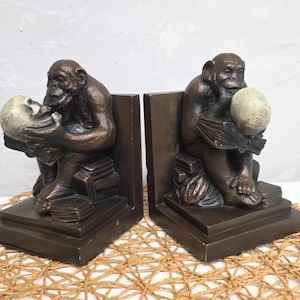 May include: Pair of bronze-colored bookends, each featuring a seated monkey figure. The monkey holds a skull and a book. The bookends are rectangular with a dark brown finish, suitable for a home library or office.