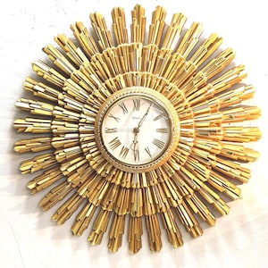 May include: A gold sunburst wall clock featuring a circular face with black Roman numerals and thin hands. The frame consists of multiple layers of radiating, pointed golden rays that extend outward from the center in a starburst pattern.