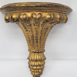 May include: Ornate gold-colored wall shelf with a decorative design. The shelf has a rounded top with a detailed edge and a stylized, fluted support. The shelf is approximately 8 inches wide.