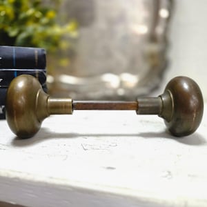 May include: A vintage brass door knob set featuring two rounded, weathered brass knobs connected by a central square metal spindle. The hardware displays a tarnished, aged patina with visible signs of wear, showcasing a classic spherical design.