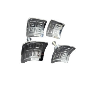 May include: Four square cabinet knobs with a polished silver finish. Each knob features a curved surface and a recessed geometric Greek key pattern divided into four quadrants. Two of the knobs include a metal mounting screw on the back side.
