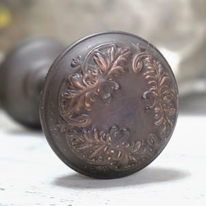 May include: A round, dark bronze-toned metal door knob featuring an embossed, raised leaf pattern around the perimeter of the face. The center of the knob is smooth and circular, with a weathered, metallic finish showing subtle copper highlights.
