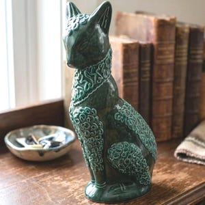 May include: A dark green ceramic cat figurine featuring a glossy finish and intricate, raised swirling patterns across its head, neck, chest, and back. The sculpture is depicted in a seated position with pointed ears and a smooth, solid base.