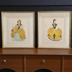 May include: Two framed prints featuring illustrations of figures in historical-style yellow dresses with polka dots, green accents, and hats. Each print is encased in a thin gold-colored frame with a cream-colored mat and multi-layered border details.