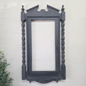 May include: A dark gray, ornate, empty picture frame. The frame has a rectangular opening, with decorative, turned posts on either side. The top of the frame has a decorative pediment with finials. The frame is painted a matte gray.