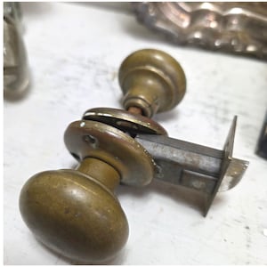 May include: A brass door knob set featuring two rounded, weathered brass knobs connected by a central metal spindle and a rectangular latch plate. The hardware shows signs of age with a dull, tarnished finish and visible mounting screw holes.