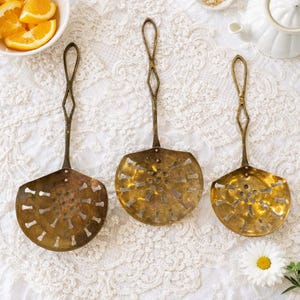 May include: Three antique brass kitchen skimmers with long handles and perforated bowls. The skimmers are arranged on a white lace tablecloth, with a bowl of orange slices and a white teapot in the background.