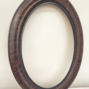May include: Oval picture frame with a dark brown, wood-grain finish. The frame has a rounded profile with a black inner edge. The frame is empty, with a white background visible through the opening. The frame is a decorative item.