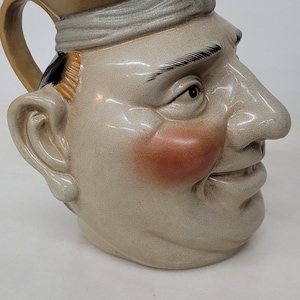 May include: A ceramic pitcher shaped like a man's head, with a tan and gray color scheme. The pitcher has a handle, a spout, and a painted face with rosy cheeks and black eyebrows. The head is wearing a hat.
