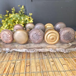 May include: A collection of antique door knobs in various metallic finishes, including bronze and brass. Some knobs feature ornate floral designs, while others have a simple, rounded shape. The knobs are arranged on a decorative silver tray.
