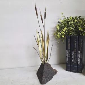 May include: A metal sculpture featuring three brown cattails and several gold-toned leaves extending from a dark, textured, triangular stone base. The sculpture stands upright, showcasing slender stems and pointed foliage in a vertical arrangement.