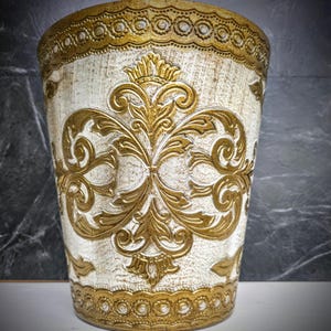 May include: A decorative wastebasket with a cream-colored base and ornate gold filigree design. The wastebasket features a repeating pattern of gold trim at the top and bottom. The central design is a symmetrical floral pattern in gold.