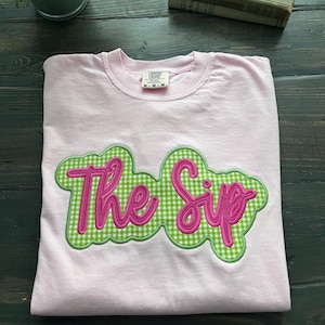May include: A light pink crewneck t-shirt featuring a large, central applique patch. The patch has a green and white gingham pattern with a scalloped edge and displays the text The Sip in bright pink, cursive, embroidered lettering.