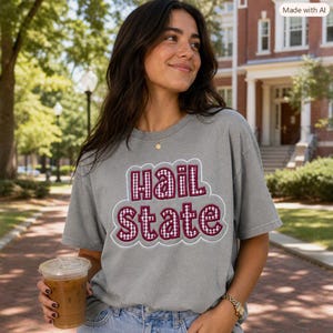 May include: A grey short-sleeve t-shirt featuring the text "hail state" in a maroon and white gingham pattern with a white outline. The shirt has a crew neckline and a relaxed fit, showcasing a casual style with a prominent graphic design.