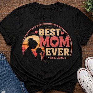 K&ouml;nnte beinhalten: Schwarzes T-Shirt mit dem Schriftzug "BEST MOM EVER" in Retro-Design. Das Design zeigt eine Mutter-Kind-Silhouette, ein Herz und "EST. 2026". Das Shirt wird mit blauen Jeans und wei&szlig;en Turnschuhen pr&auml;sentiert.