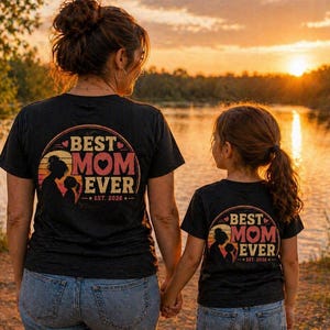 K&ouml;nnte beinhalten: Zwei schwarze T-Shirts mit dem Aufdruck "BEST MOM EVER" in einem kreisf&ouml;rmigen Design. Das Design enth&auml;lt eine Silhouette einer Mutter und eines Kindes. Die Shirts werden von einer Mutter und ihrem Kind an einem See bei Sonnenuntergang getragen.