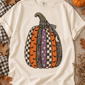 May include: An off-white short-sleeve t-shirt featuring a large graphic of a pumpkin composed of vertical panels with various patterns including orange with black bats, black with spiderwebs, purple with white stars, and black and white checkers.