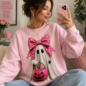 Op de afbeelding: Een lichtroze sweater met ronde hals en een afbeelding van een witte geest met een grote roze strik met stippen. De geest houdt een felroze pompoenemmer vol snoepjes vast en er is een kleine zwarte spin aan de zijkant zichtbaar.