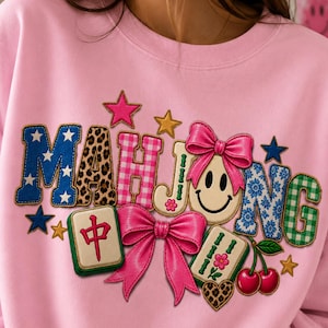 May include: A light pink sweatshirt featuring the word MAHJONG in colorful, patterned letters. The design includes a smiley face with a pink bow, two mahjong tiles, a pair of red cherries, a leopard print heart, and several small stars.