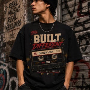 May include: Black t-shirt with the text "STILL BUILT DIFFERENT" and a cassette tape graphic that reads "GOLDEN ERA" and "90". The shirt also has the text "NO SKIP. NO ALGORITHM." The person is wearing a black beanie and a silver chain.