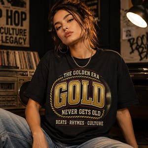 Op de afbeelding: Een zwart t-shirt met korte mouwen en een grafische print met de tekst THE GOLDEN ERA GOLD NEVER GETS OLD BEATS RHYMES CULTURE. Het ontwerp bevat een goudkleurige touwrand en een afbeelding van een vinylplaat op de borst.