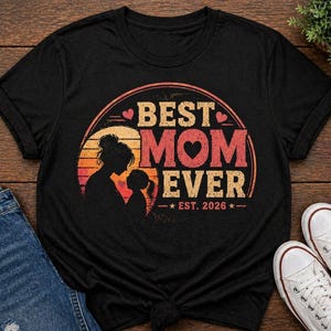 K&ouml;nnte beinhalten: Schwarzes T-Shirt mit dem Aufdruck "BEST MOM EVER" in Retro-Design. Das Design zeigt eine Mutter-Kind-Silhouette, ein Herz und den Text "EST. 2026". Das Shirt wird mit blauen Jeans und wei&szlig;en Turnschuhen pr&auml;sentiert.