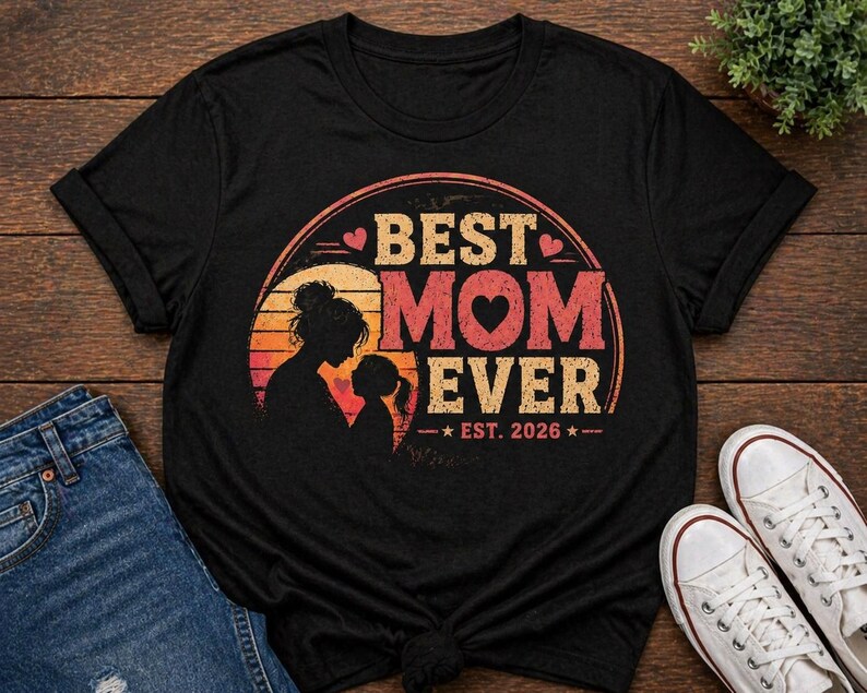 K&ouml;nnte beinhalten: Schwarzes T-Shirt mit dem Aufdruck "BEST MOM EVER" in Retro-Design. Das Design zeigt eine Mutter-Kind-Silhouette, ein Herz und den Text "EST. 2026". Das Shirt wird mit blauen Jeans und wei&szlig;en Turnschuhen pr&auml;sentiert.