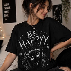 Può includere: T-shirt nera con la scritta "BE HAPPY" in un font invecchiato, con un mostro dei cartoni animati con grandi occhi e braccia alzate. La maglietta ha un girocollo e maniche corte.