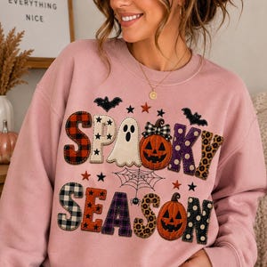 Op de afbeelding: Een oudroze sweater met ronde hals en de tekst SPOOKY SEASON in kleurrijke letters met verschillende patronen. Het ontwerp bevat een wit spookje, twee oranje pompoenen, een spinnenweb, een kleine spin, zwarte vleermuizen en kleine sterren.