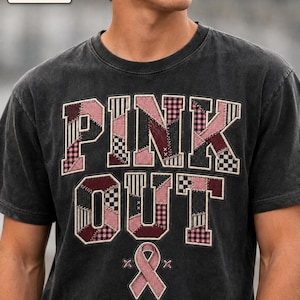 May include: A charcoal grey short-sleeve t-shirt featuring the text PINK OUT in a patchwork style with various shades of pink, maroon, and black and white patterns. Below the text is a pink awareness ribbon flanked by two small cross stitch symbols.