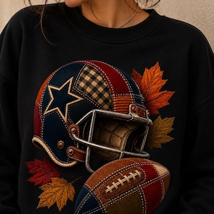 Könnte beinhalten: Ein schwarzes Sweatshirt mit einer Grafik eines Football-Helms und eines Footballs, beide im Patchwork-Design mit blauen, roten und braunen Stofftexturen. Der Helm zeigt einen Stern und der Football hat weiße Schnürsenkel, umgeben von Herbstblättern.