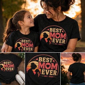 K&ouml;nnte beinhalten: Schwarze T-Shirts mit einem kreisf&ouml;rmigen Aufdruck &bdquo;BEST MOM EVER&ldquo; in Rosa und Wei&szlig;, mit einer Silhouette von Mutter und Kind. Der Text &bdquo;EST. 2026&ldquo; ist ebenfalls enthalten. Die Shirts werden von einer Mutter und ihrer Tochter getragen.