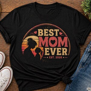 K&ouml;nnte beinhalten: Schwarzes T-Shirt mit Retro-Design, das "BEST MOM EVER" in orangefarbenen und roten Buchstaben zeigt. Das Design beinhaltet eine Mutter-Kind-Silhouette, mit dem Jahr "EST. 2026" darunter.